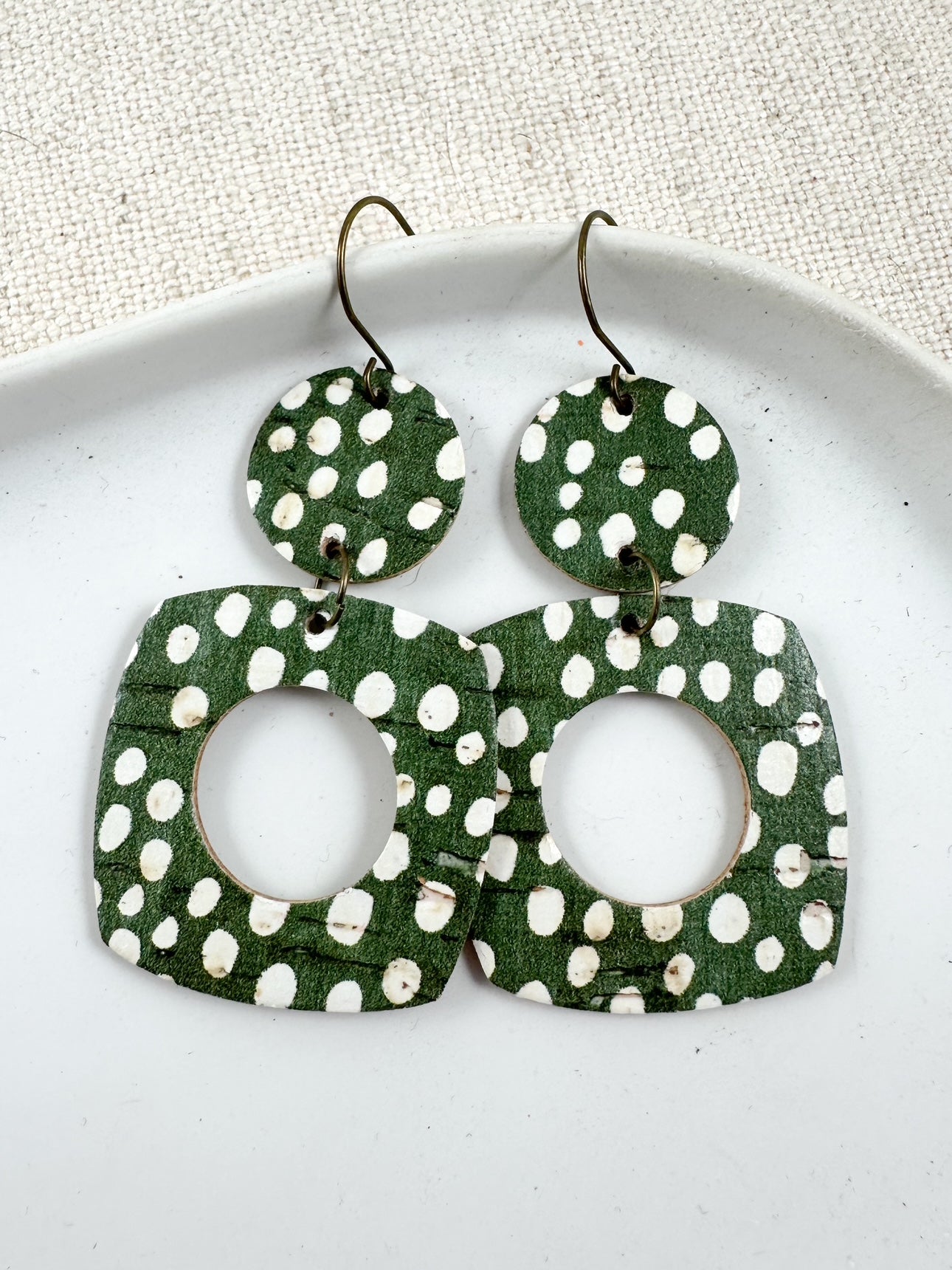Moss Meadow Large Birch cork earrings displayed on a white dish, featuring a deep moss green background with ivory dots and antique brass hooks.