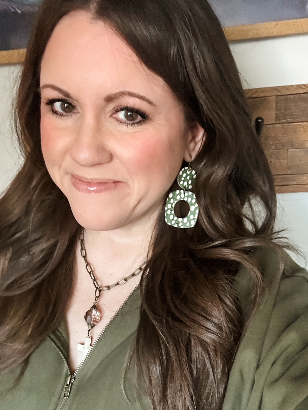 Woman wearing Moss Meadow Large Birch cork earrings with white polka dots and an olive green base, paired with a green zip-up top and bronze cross necklace.