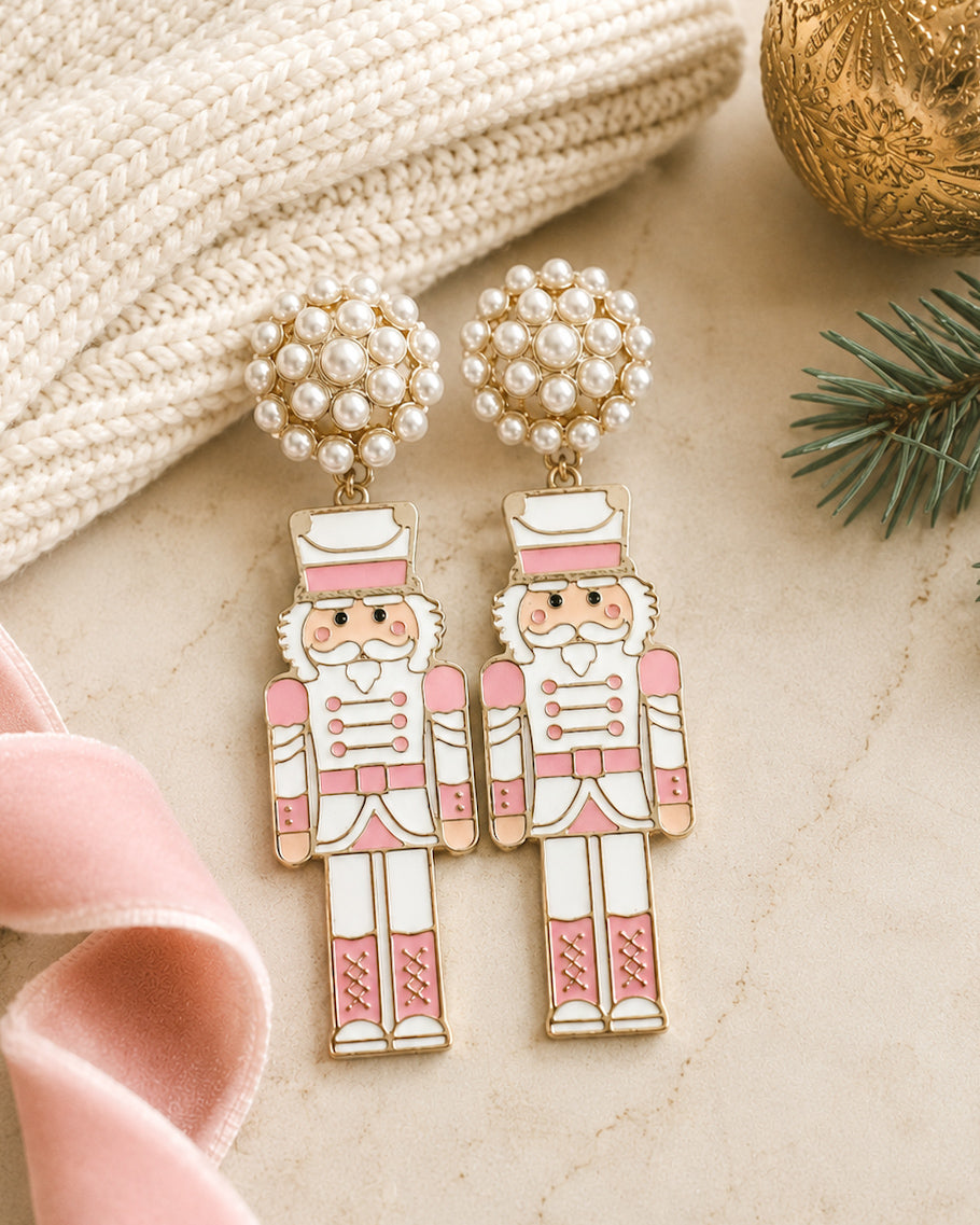 Blush pink nutcracker earrings styled with a cream knit sweater, blush velvet ribbon, and holiday greenery.