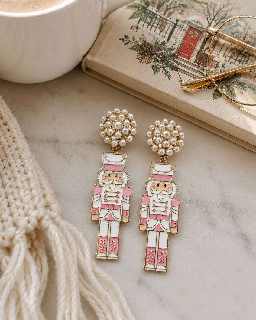 Blush pink nutcracker earrings styled beside a holiday book, coffee, and cozy winter accessories.