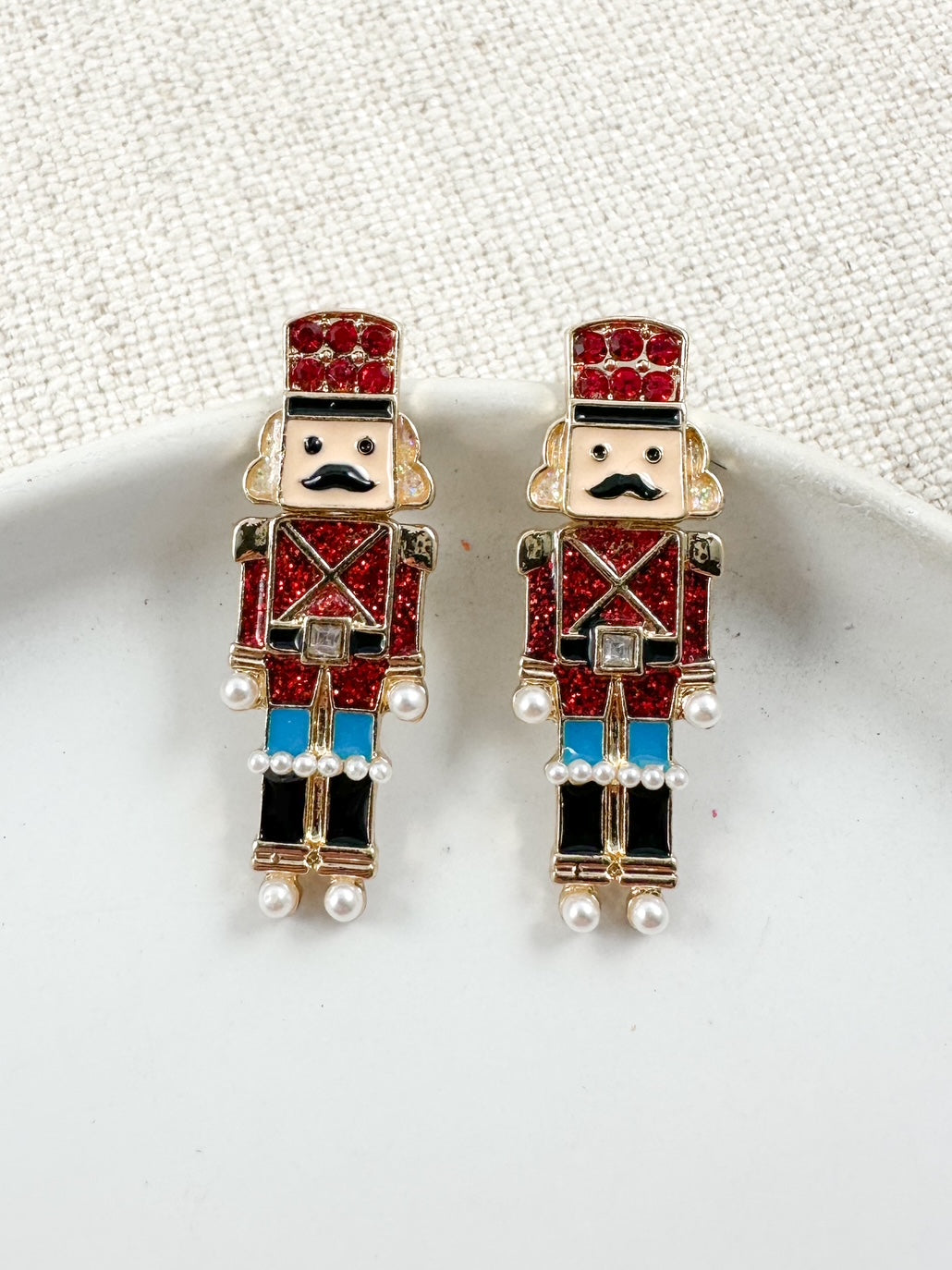 Red Nutcracker soldier earrings with glittered uniforms and small pearl details displayed on a white dish.