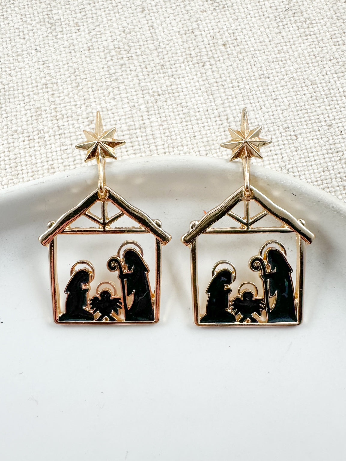 Black and gold nativity scene earrings featuring Mary, Joseph, and baby Jesus under a star, displayed on a white dish.