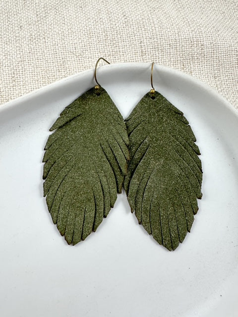 Close-up of long olive green suede feather-shaped earrings on fishhook ear wires, displayed on a white dish.