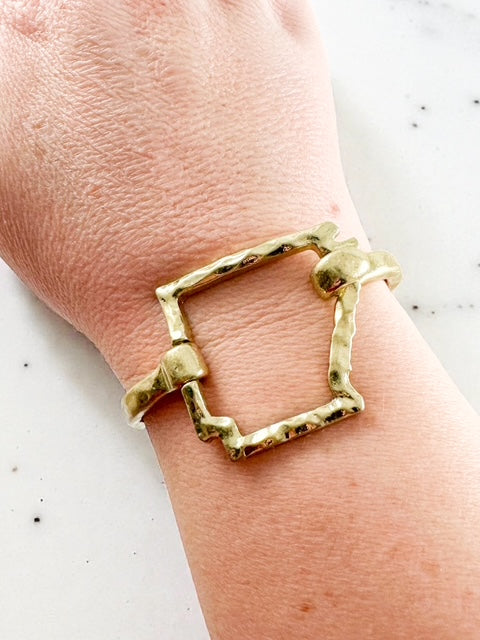 Worn gold hammered Arkansas-shaped cuff bracelet shown on a wrist against a light surface.