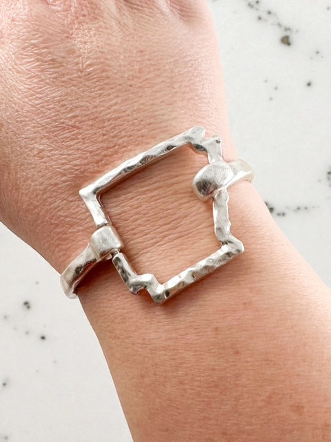 Worn silver hammered outline cuff bracelet shown on wrist against a light surface.