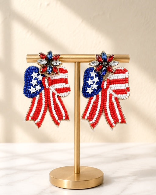 Patriotic Bow Earrings with red, white, and blue seed bead detailing and rhinestone floral posts displayed on a gold jewelry stand.