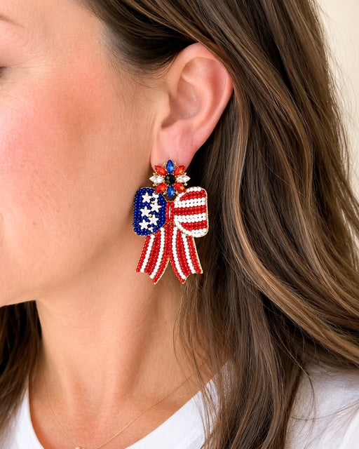 Woman wearing Patriotic Bow Earrings featuring red, white, and blue seed bead bows with sparkling rhinestone accents.