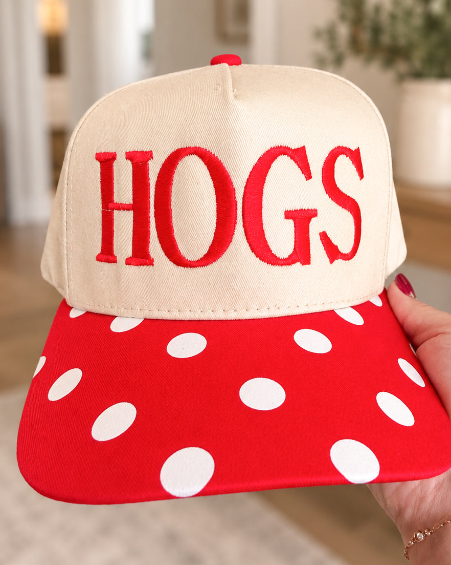 Close-up of a cream HOGS hat held in one hand, showing the red embroidered lettering, cream fabric, and red bill with white polka dots.