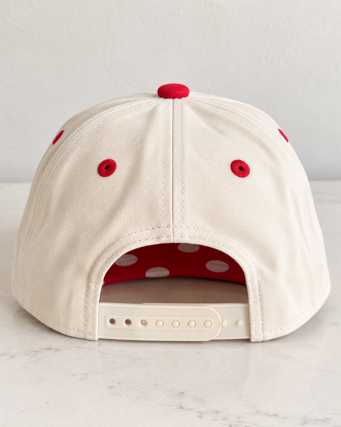 Back view of the cream Pep Rally Hat showing the red top button, red embroidered eyelets, and adjustable cream snapback closure.