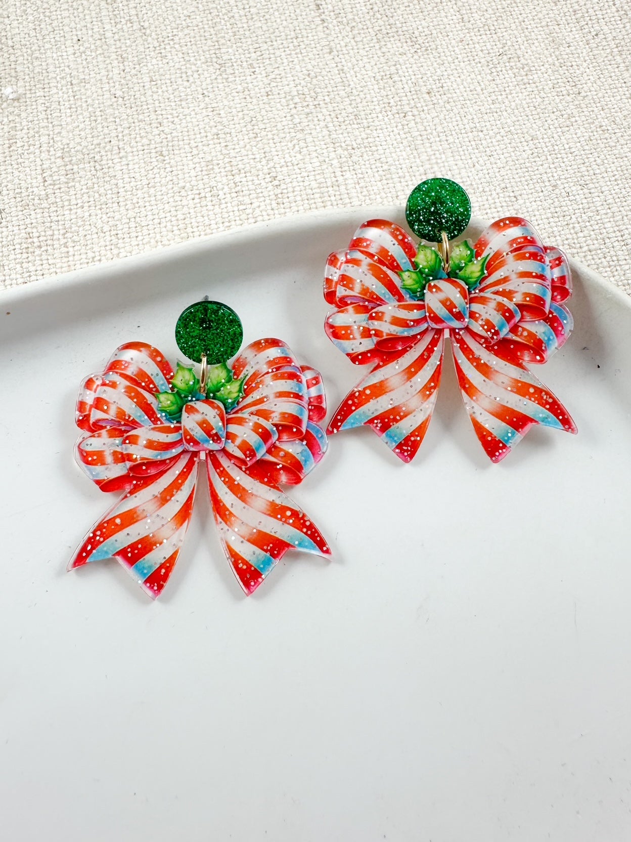Red and white peppermint-striped bow earrings with green glitter studs displayed on a white dish.
