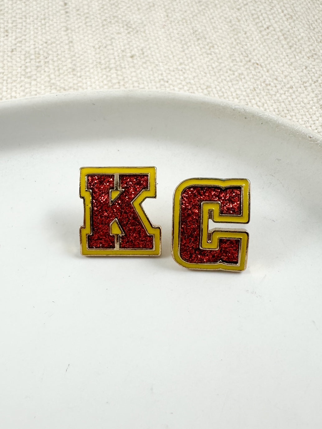 Red glitter letter K and C stud earrings with gold and yellow trim displayed on a white dish