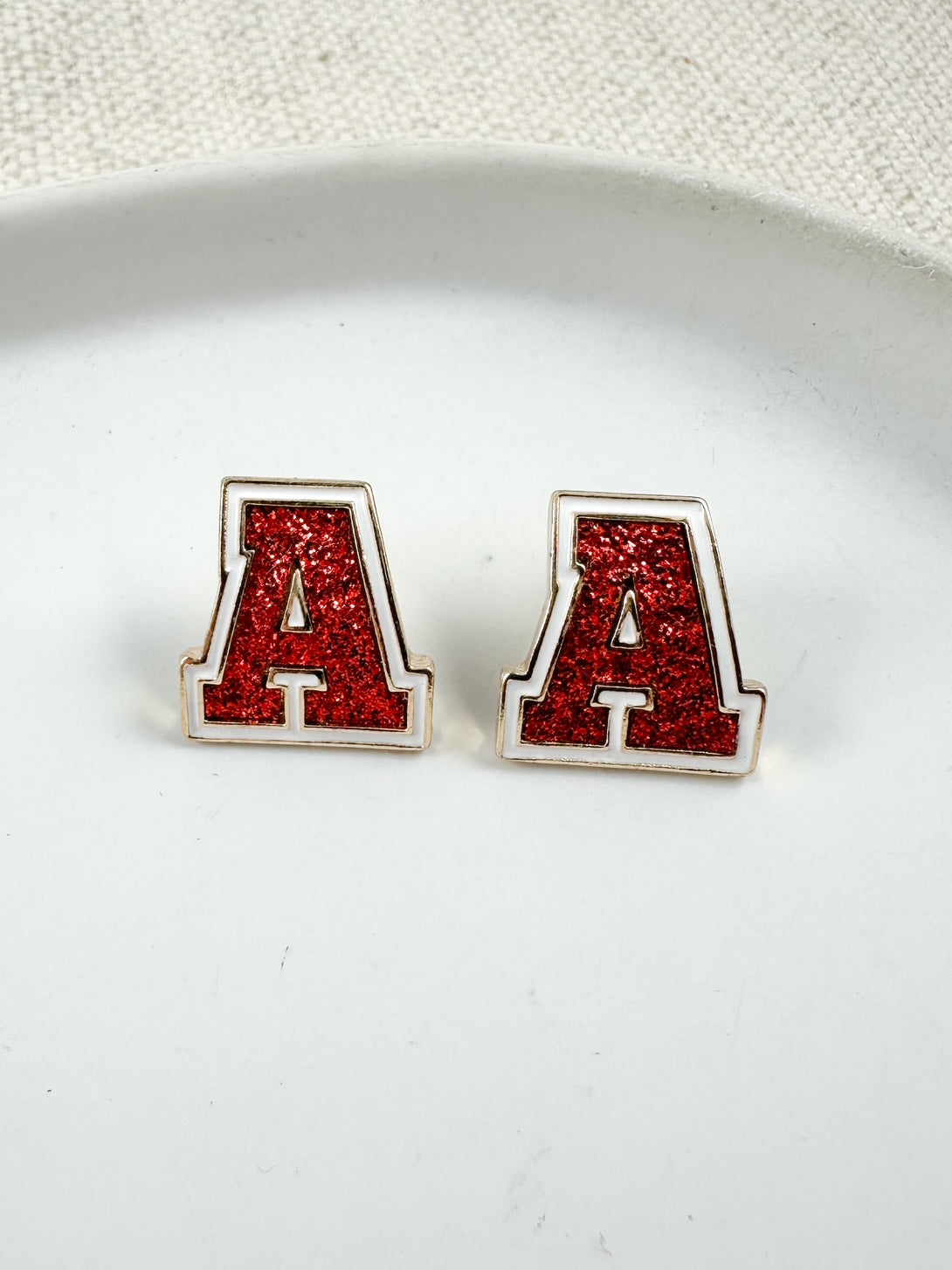 Red glitter letter A stud earrings with gold trim displayed on a white dish