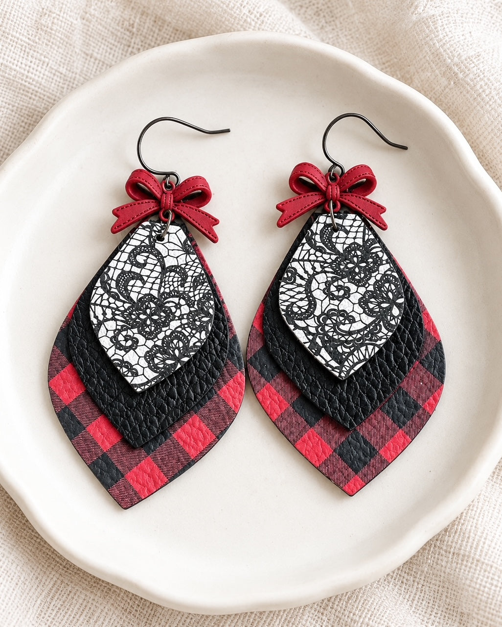 Pretty Little Rebel Earrings displayed on an ivory ceramic dish, showing layered red buffalo plaid, black textured leather, lace-print details, and red bows.