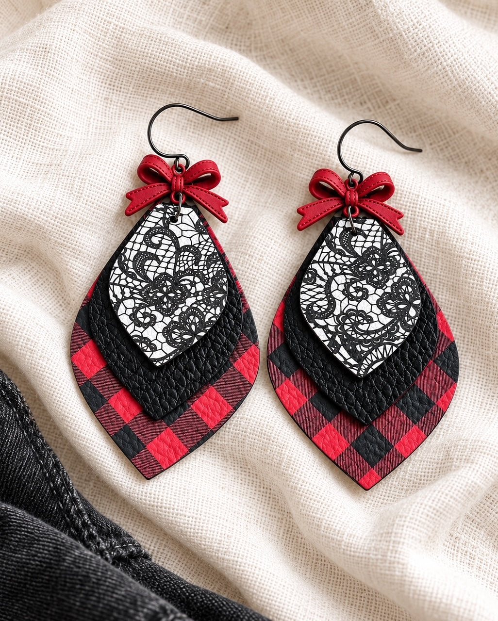 Handmade red-and-black buffalo plaid earrings with black leather layers, floral lace print, and red bow accents styled on soft ivory linen fabric.
