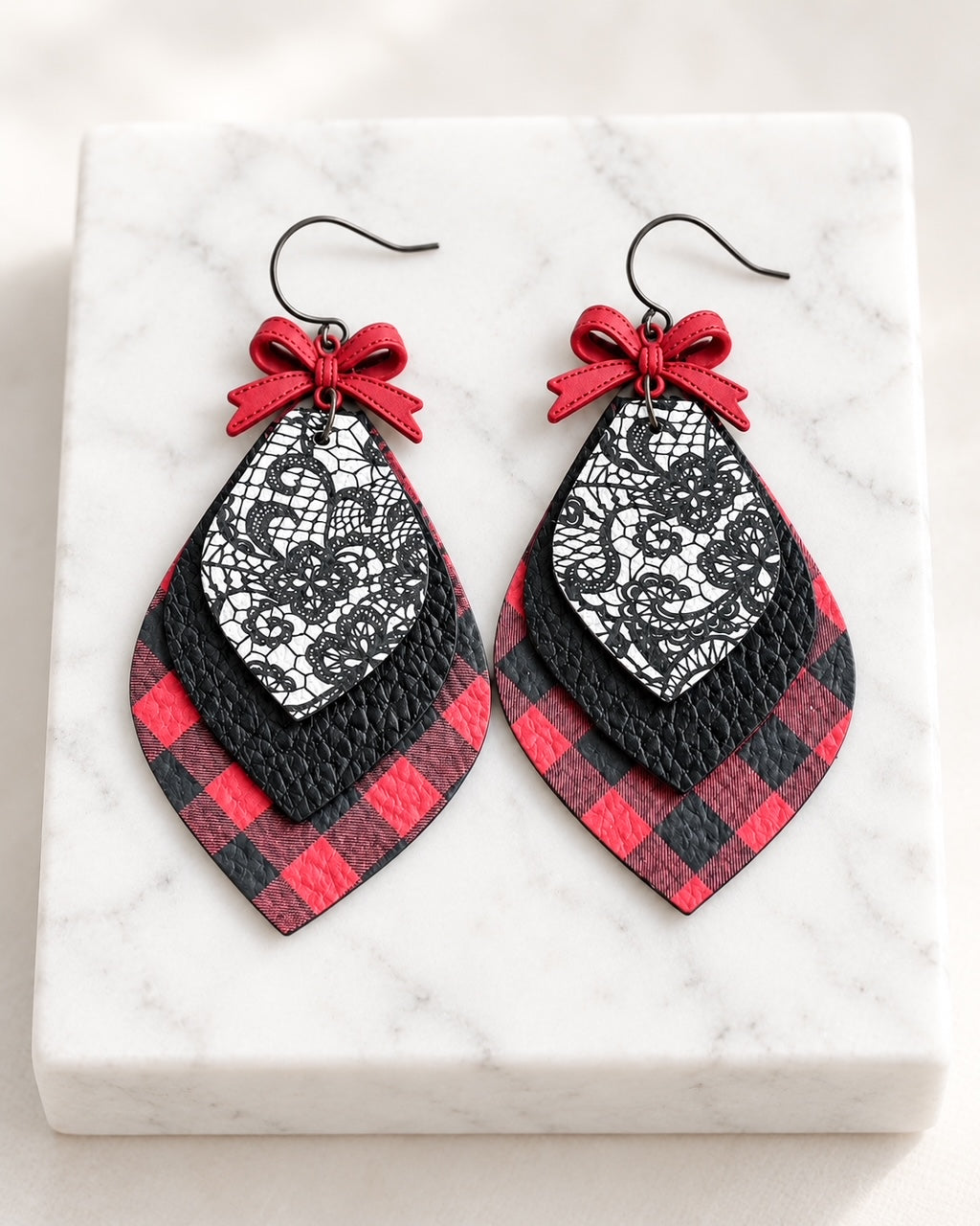 Pretty Little Rebel handmade leather earrings on white marble with red-and-black buffalo plaid, black textured leather, delicate lace pattern, and dimensional red bows.