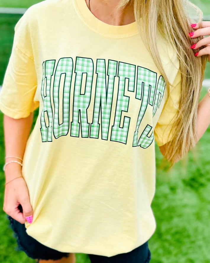 Woman wearing a light yellow tee with oversized HORNETS lettering in green and white gingham.