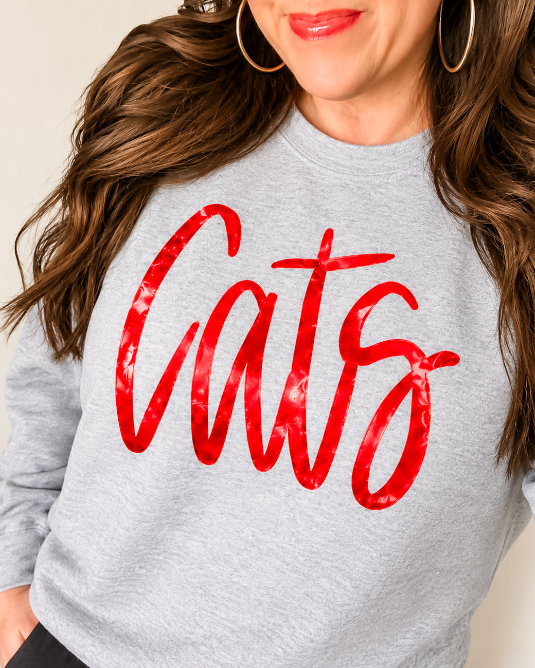 Woman wearing a sport gray Cats crewneck sweatshirt with shiny metallic red script lettering and gold hoop earrings.
