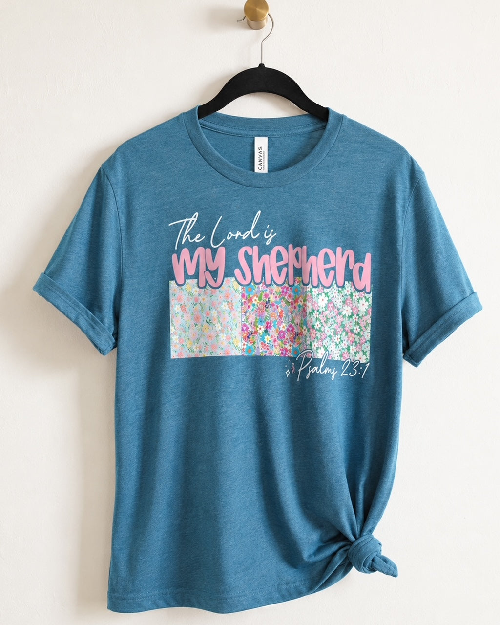 Shepherd's Garden Tee hanging on a wooden hanger showing the floral Psalm 23 "The Lord Is My Shepherd" design.