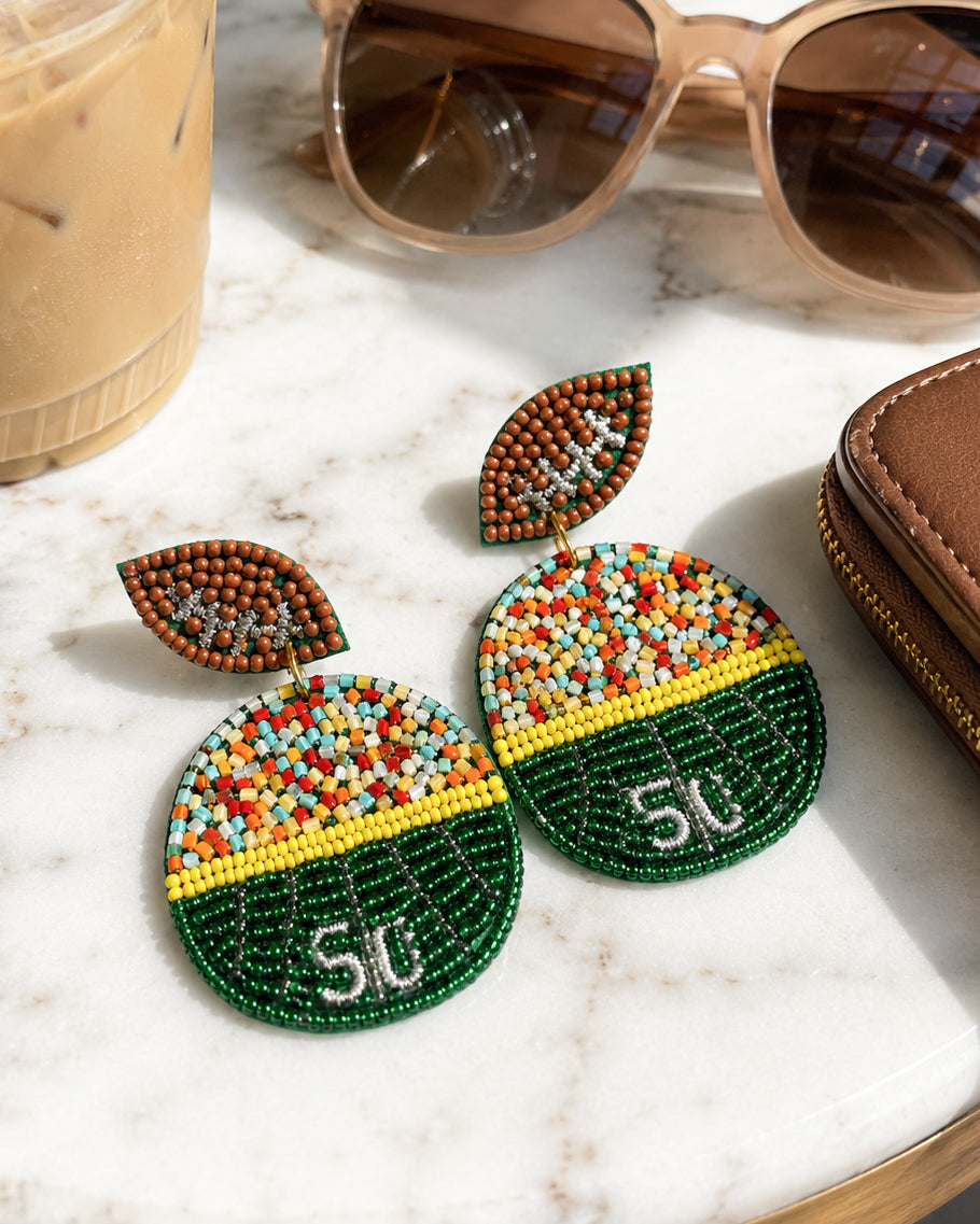 Green football field seed bead statement earrings on white marble beside sunglasses, coffee, and a brown wallet.