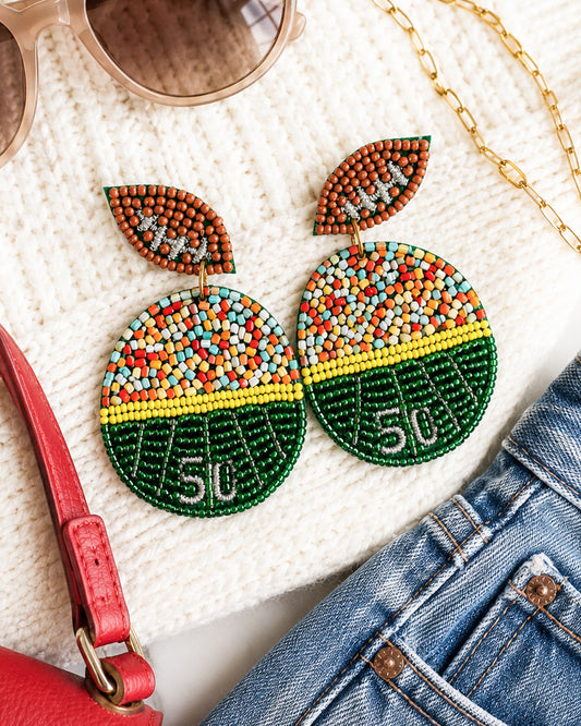 Football field seed bead earrings styled on a cream sweater with denim, sunglasses, and gold jewelry.