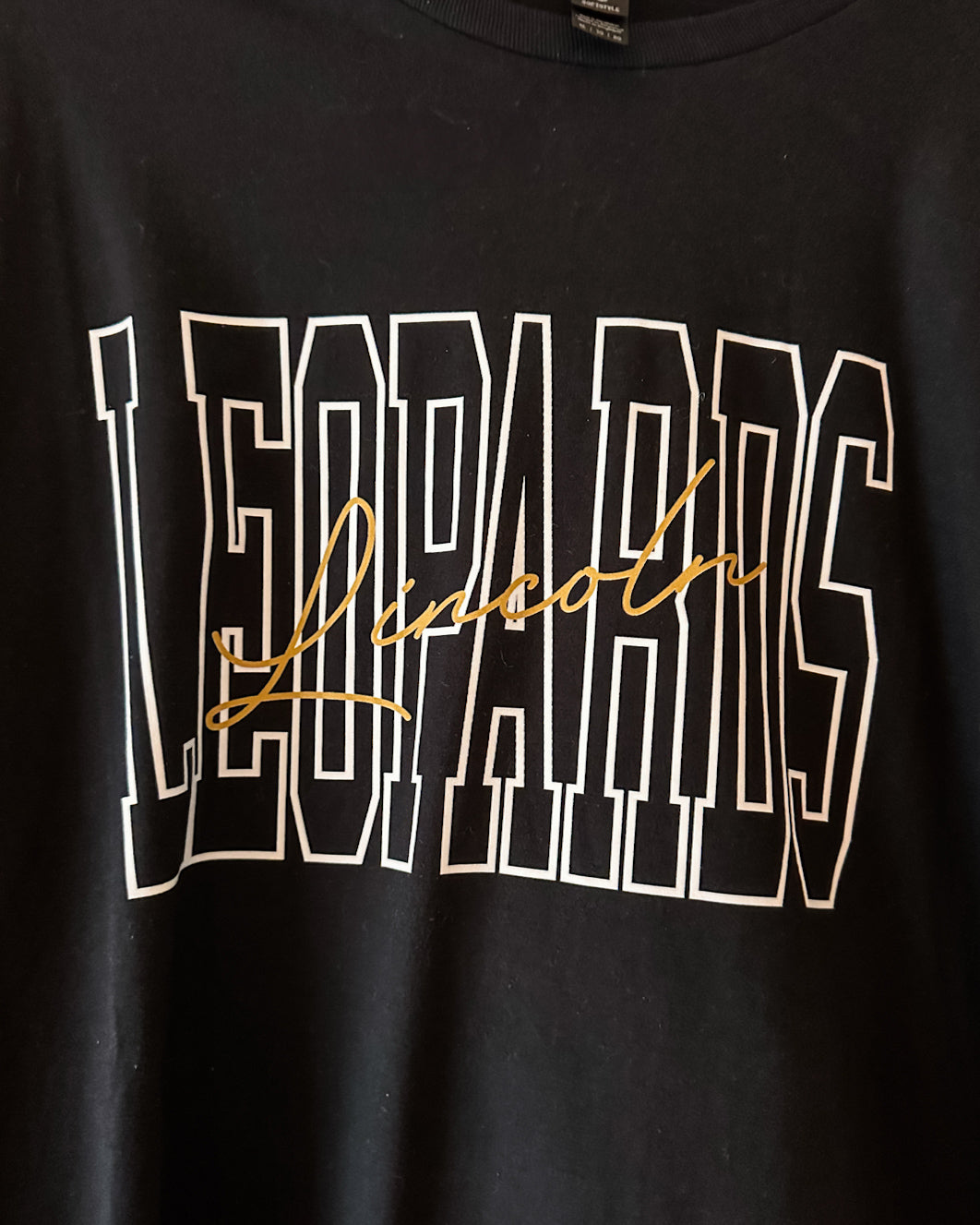 Close-up of black Lincoln Leopards shirt featuring white outlined varsity lettering with gold Lincoln script across the front.