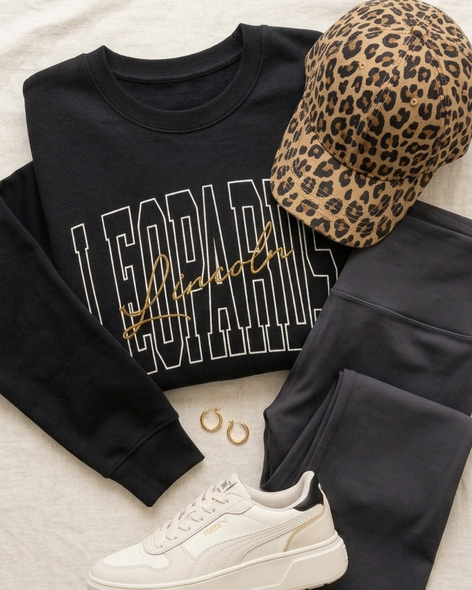 Black Lincoln Leopards graphic tee styled with medium-wash jeans, white sneakers, black crossbody bag, and gold bracelets.