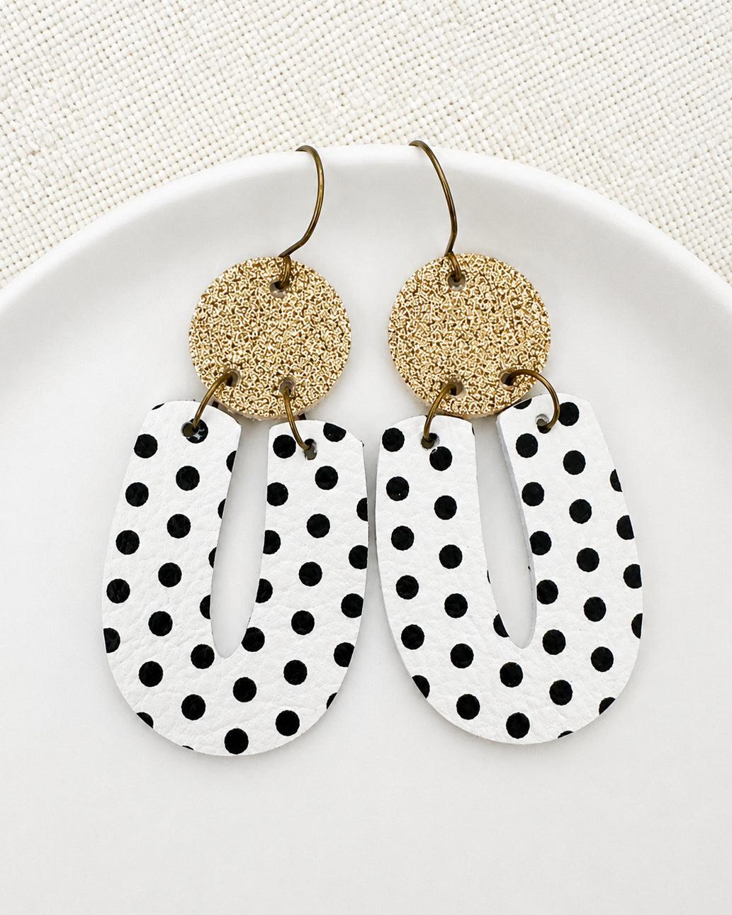 Black and white polka dot earrings with matte gold glitter tops styled on a small white dish against a soft neutral background.
