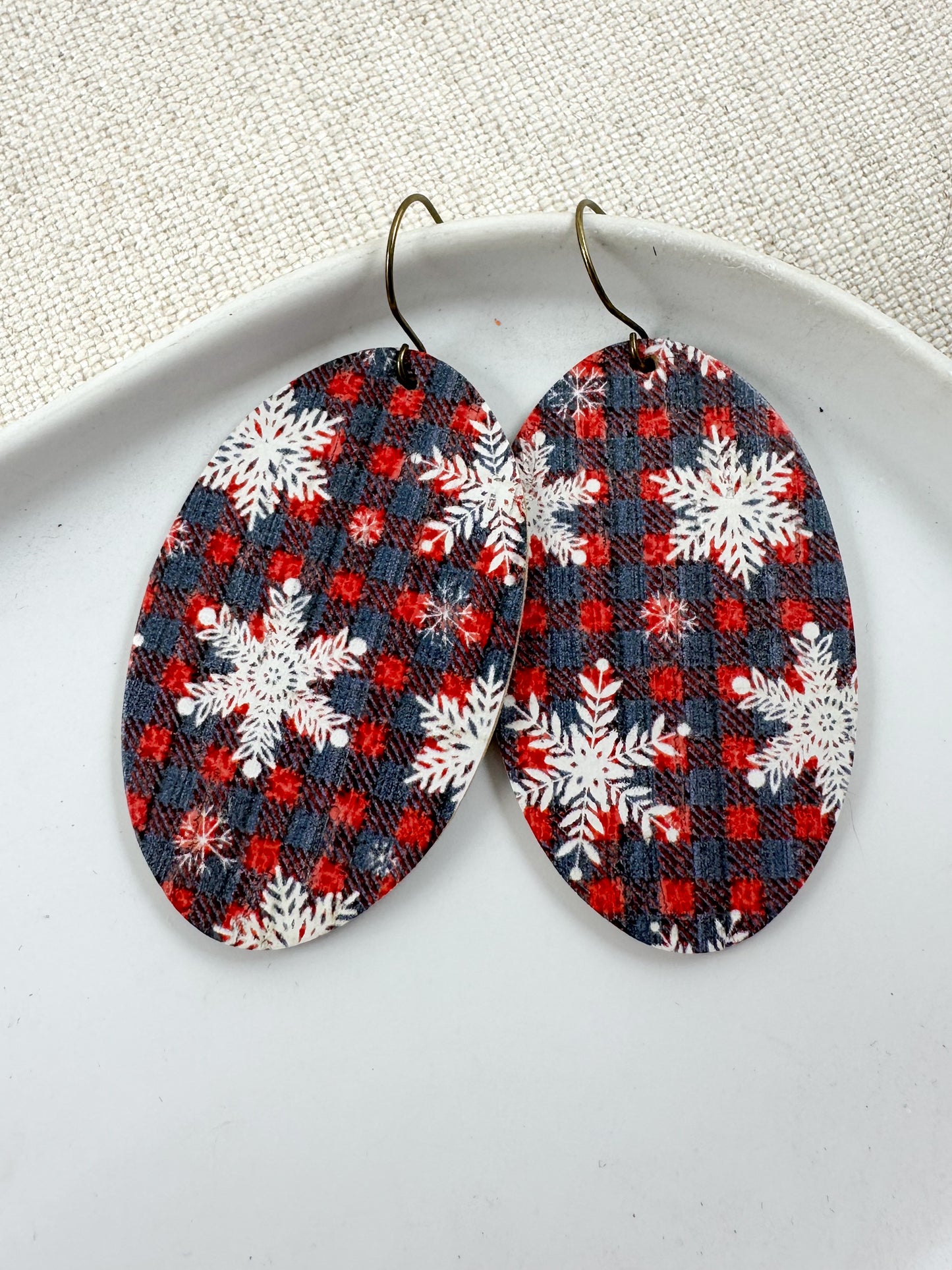 Close-up of large oval Snowflake & Flannel Cork dangle earrings with red and navy plaid and white snowflakes on a white dish.