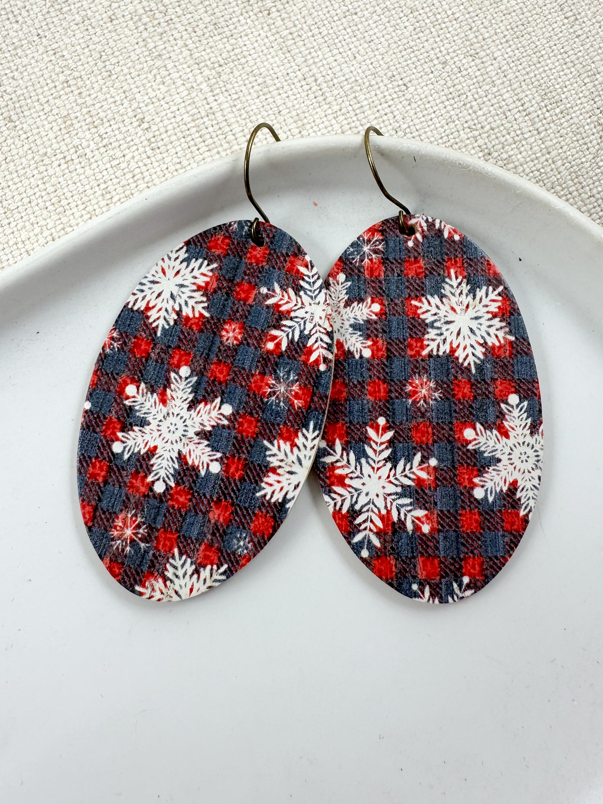 Close-up of large oval Snowflake & Flannel Cork dangle earrings with red and navy plaid and white snowflakes on a white dish.