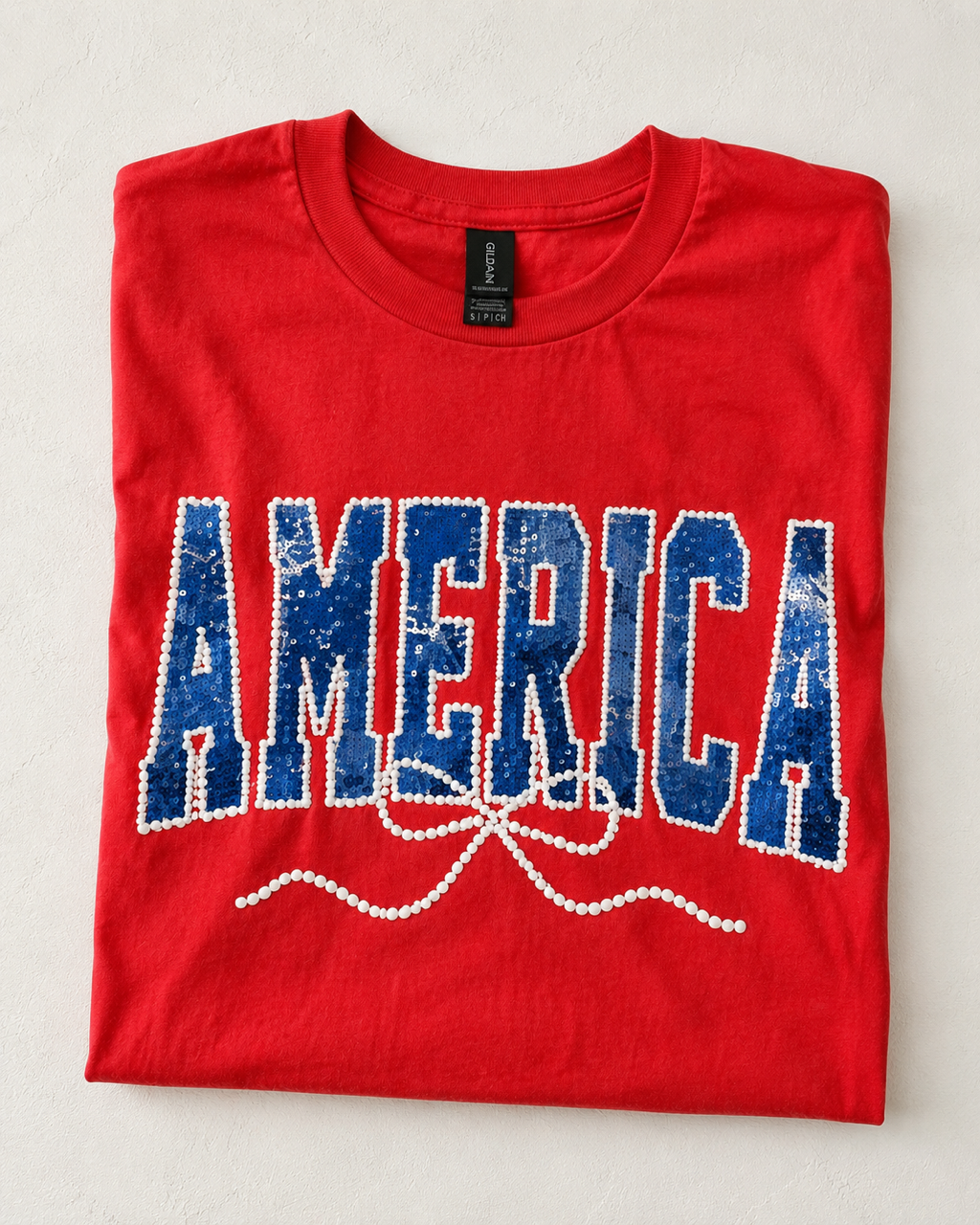 Folded Sparkler Season Tee with faux sequin America graphic and pearl-inspired accents on a red Gildan unisex tee.