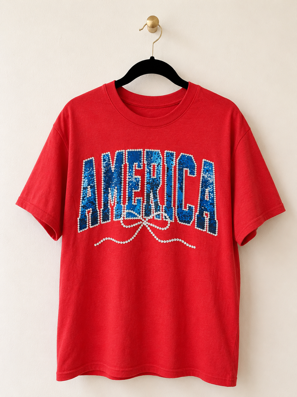 Sparkler Season Tee hanging on a black velvet hanger featuring a faux sequin America graphic on a red unisex tee.