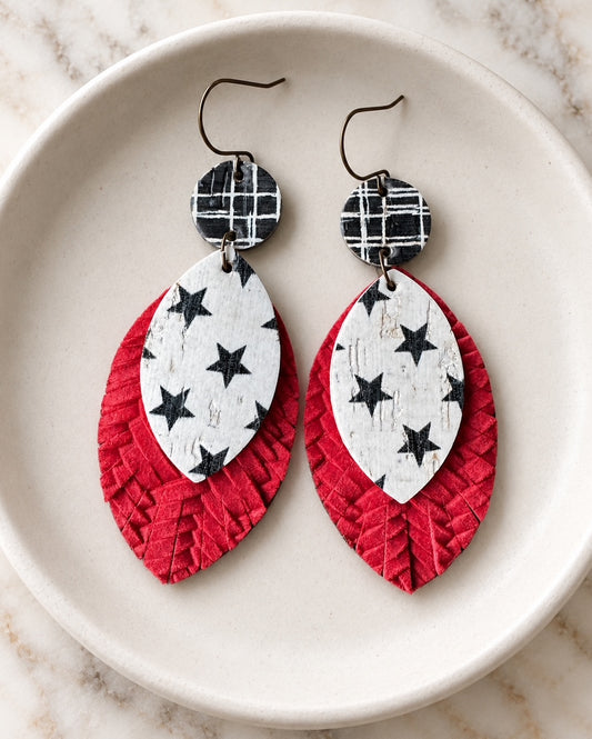 Pair of handmade Starstruck Earrings displayed on a cream ceramic dish, featuring black-and-white plaid tops, distressed white star-print layers, and textured red leather.