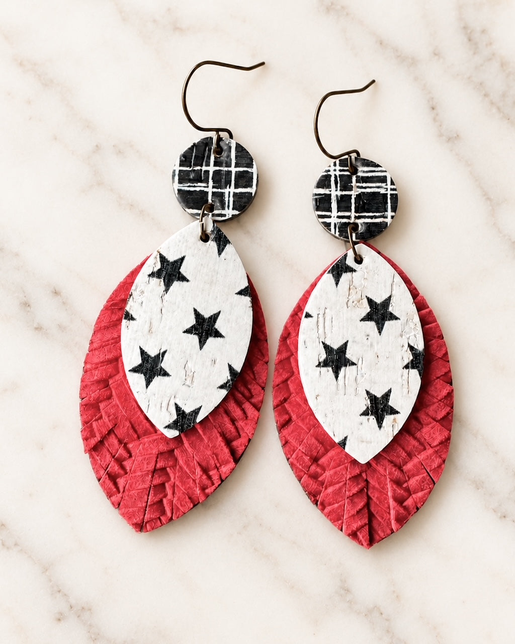 Close-up of Starstruck layered leather and cork earrings on white marble, showing black plaid circle tops, white teardrops with black stars, and textured red leather backing.