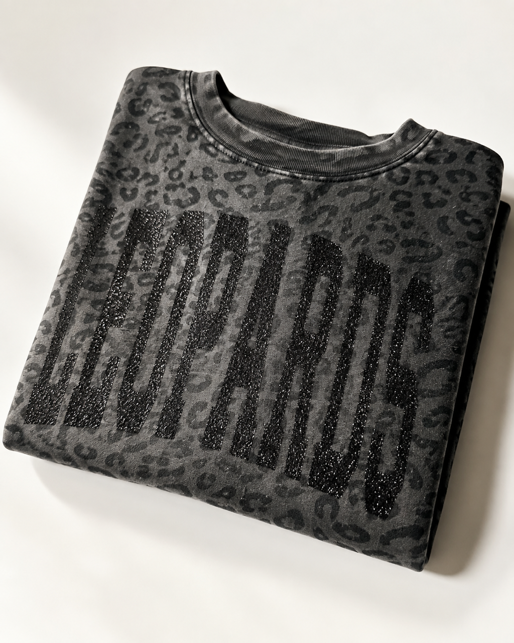 Folded black leopard print LEOPARDS graphic tee showing the black glitter varsity lettering.