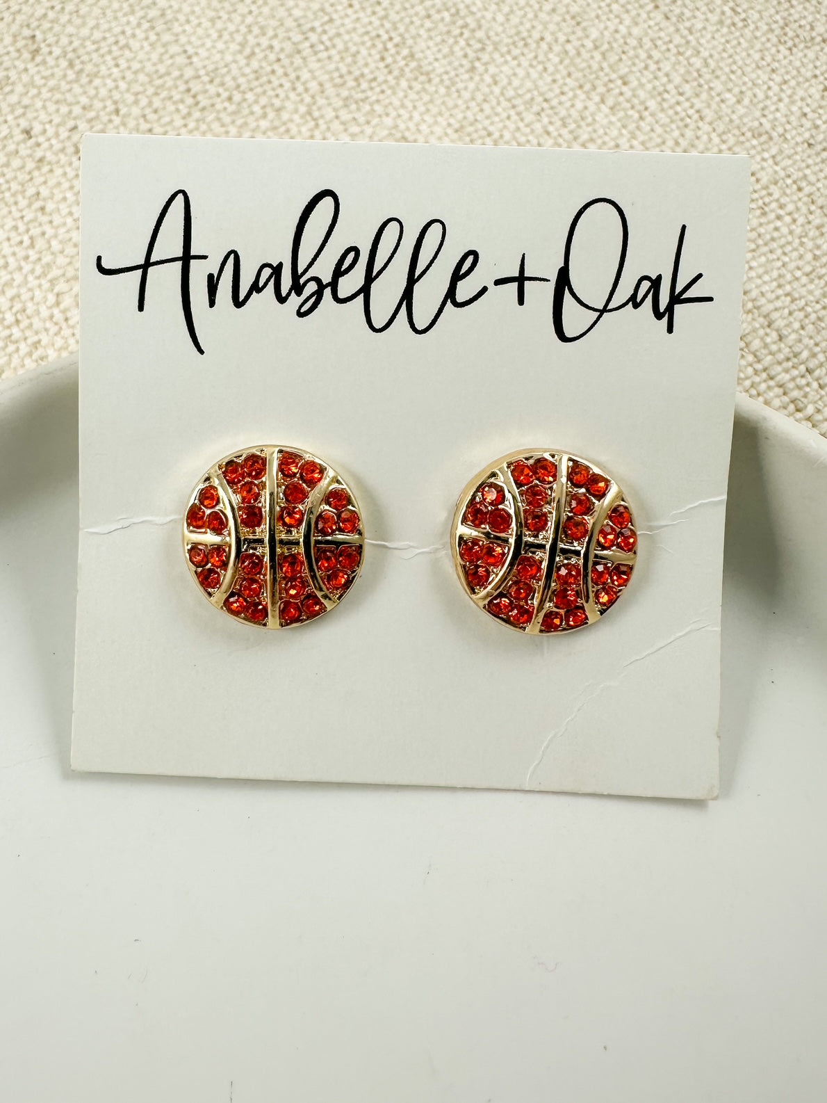 Gold-tone basketball-themed stud earrings with small crystal accents displayed on a white dish