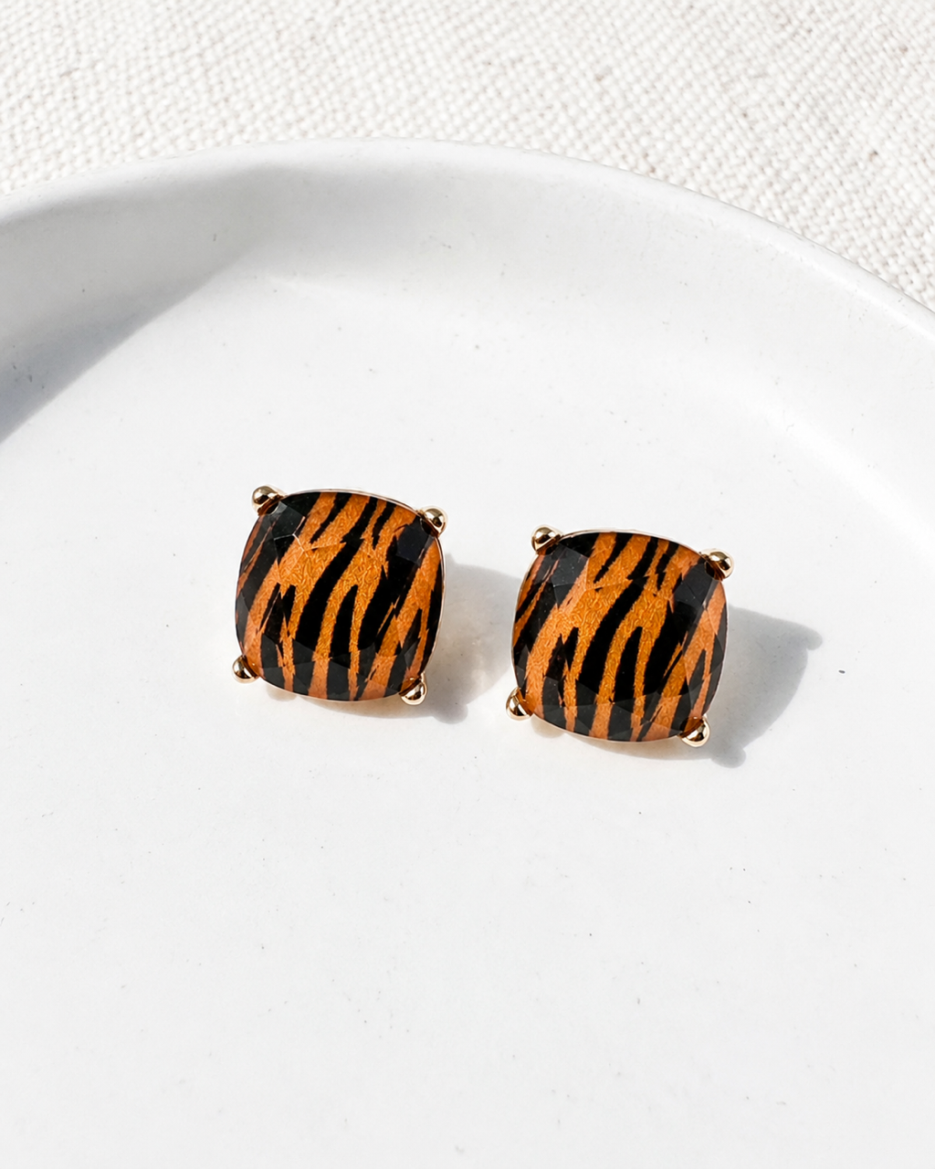 Tiger Trail tiger print stud earrings with faceted cushion-cut stones and gold-tone settings on a white ceramic dish.