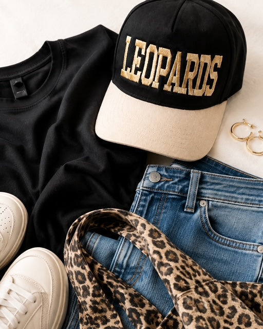 Varsity Leopard Hat styled in a flat lay with a black top, medium-wash jeans, cream sneakers, gold hoops, and a leopard-print bag. The black-and-ivory snapback hat features dimensional embroidered LEOPARDS lettering across the front.