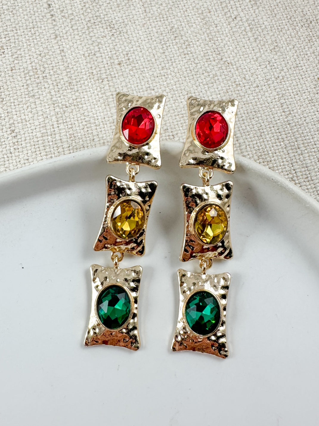 Long gold-tone drop earrings with three stacked rectangular frames each holding faceted oval stones in red, gold, and green displayed on a white dish