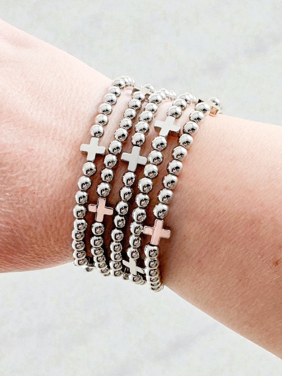 Five silver tone stretch beaded bracelets with cross station accents stacked together on a woman’s wrist.