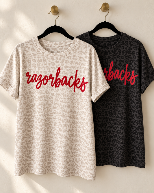 Two leopard print graphic tees hanging on a neutral wall, featuring custom script lettering across the front.