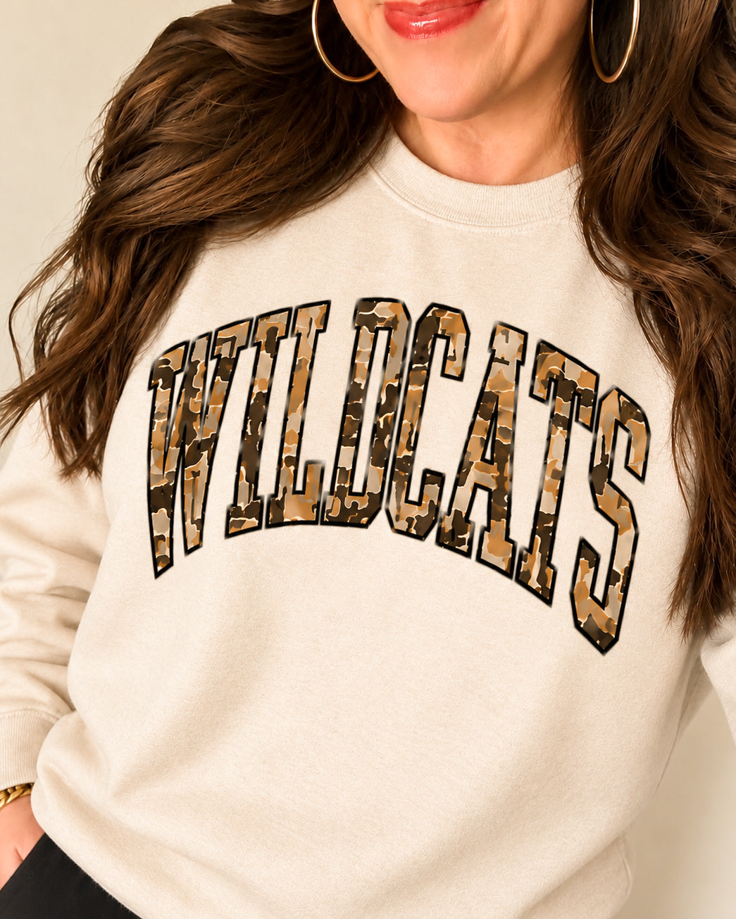 Woman wearing the sand Wildcat Territory sweatshirt with camouflage WILDCATS lettering, black pants, and gold hoops.