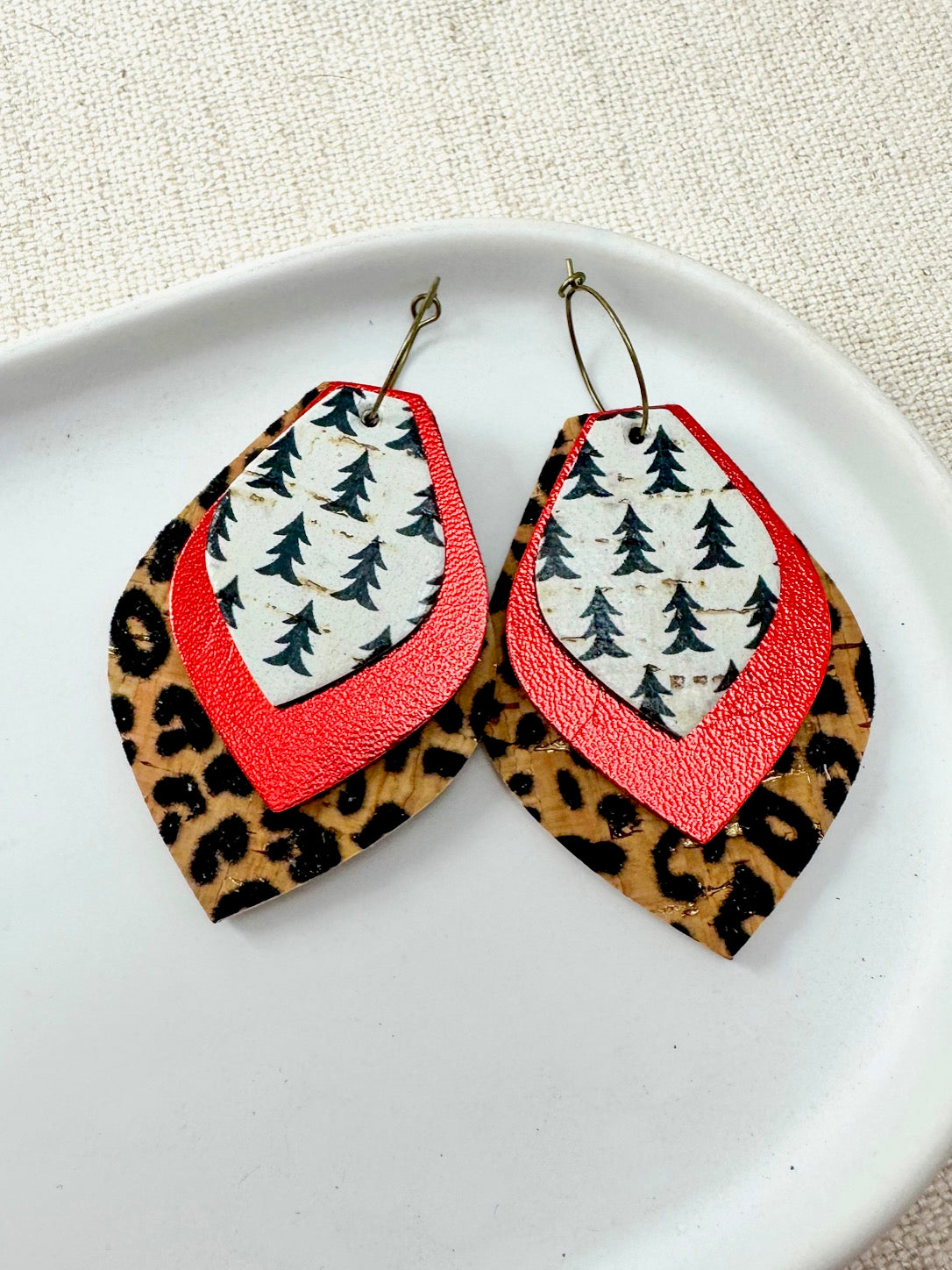 Close-up of Wildwood Noel Earrings featuring layered leopard print, red shimmer, and pine tree cork on antique brass hoops, part of the Mix & Match Collection.